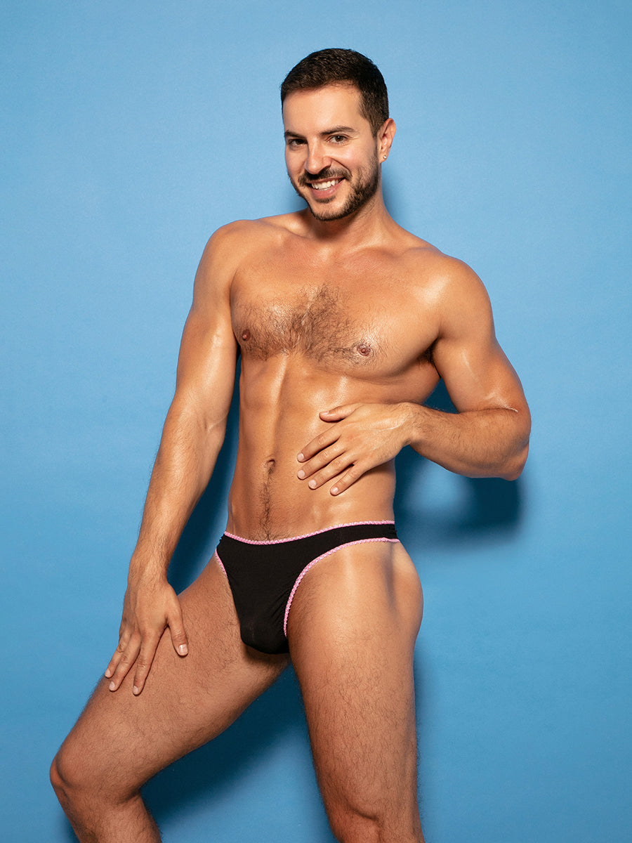 Front view of a man wearing black thong panties with pink lace trim, against a blue background.
